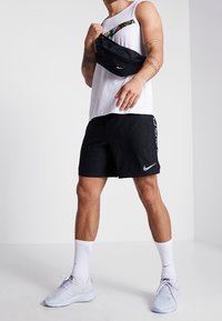 Bermudas deportivas negras con el logo de Nike, camiseta sin mangas blanca, riñonera negra y zapatillas de color claro. Presenta calcetines y tatuajes visibles.