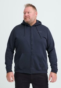 Felpa con zip di colore blu navy, dotata di tasca a marsupio, cappuccio regolabile con cordini e polsini a coste. Il materiale sembra morbido e dalla trama evidente. Logo sul petto.