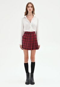 Gonna plissettata a quadri rossi con dettagli a tasca, abbinata a una camicia bianca con bottoni e calze nere fino al ginocchio con stivali.