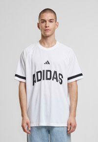Bela kratka majica s kratkimi rokavi z črnim logotipom Adidas in horizontalnimi črnimi črtami na rokavih. Narejena iz zračnega materiala, ohlapen kroj.