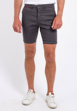 Homme portant un short gris foncé à ourlet roulé, des baskets blanches et une chemise blanche, debout devant un fond uni.