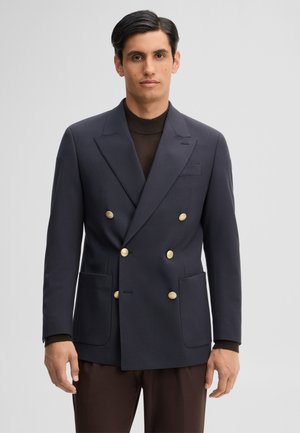 Uomo che indossa una giacca doppiopetto blu scuro con bottoni dorati e pantaloni marrone scuro, in piedi davanti a uno sfondo chiaro uniforme.