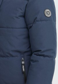 Mörkblå puffjacka med kviltat foder och synlig dragkedja. Har en rund logopatch på axeln.