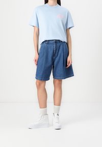 Vaaleansininen puuvillapaita, jossa on vaaleanpunainen graafinen kuvio rinnassa, yhdistettynä löysiin farkkushortseihin ja valkoisiin korkeisiin tennareihin.