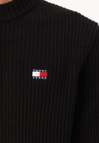 Juodas juostuotas megztinis su aukštu kaklu. Savybė - mažas išsiuvinėtas logotipas raudonos, baltos ir mėlynos spalvų kairėje pusėje.