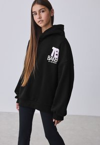 Felpa nera oversize con logo "TB" ricamato in rosa e testo "BAKER BY TED BAKER". Tessuto morbido e vestibilità rilassata, abbinata a pantaloni neri.