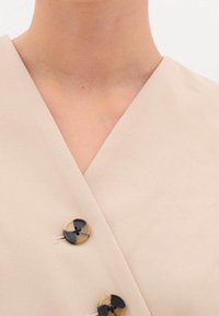 Krémový top s hlubokým výstřihem do V, zdobený dvěma dekorativními knoflíky v černobéžovém mramorovém vzoru. Hladká textilie.