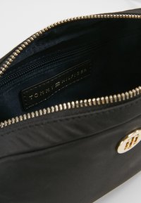 Interno di una borsa tote nera in nylon con una cerniera dorata metallizzata, una tasca con zip e un'etichetta logo in pelle nera. Texture liscia, forma strutturata.