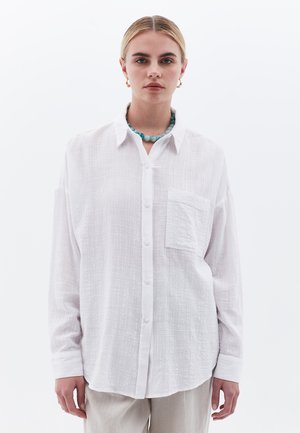 Camicia bianca a maniche lunghe con un tessuto texture, tasca frontale e orlo arrotondato. Indossata con una collana di perline blu.