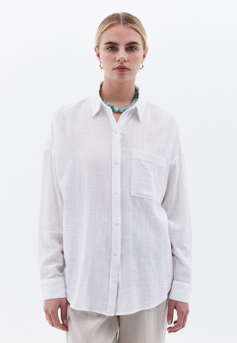 Camisa blanca de botones con una tela texturizada, de mangas largas, un bolsillo en el frente y un dobladillo redondeado. Se lleva con un collar de cuentas azules.