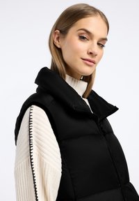 Czarna kamizelka puchowa z wysokim kołnierzem, zapięciem na zamek i bocznymi kieszeniami. Nosi się ją na kremowym swetrze z prążkowanym golfem z ciemnymi przeszyciami.