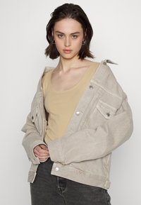 Casaco de ganga bege com grandes bolsos e puxadores prateados, usado sobre uma t-shirt sem mangas cor beige claro, combinado com jeans cinza escuro.