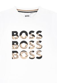 T-shirt bianca in cotone con numerosi loghi "BOSS" in nero e beige, con collo rotondo e due bottoni a pressione sulla spalla.