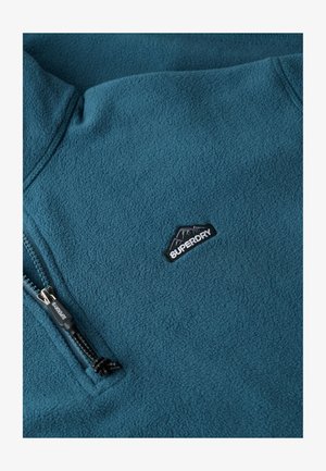 Zielona polarowa kurtka z pół zamkiem, gładką fakturą oraz czarnym haftowanym logo z motywem góry na piersi.