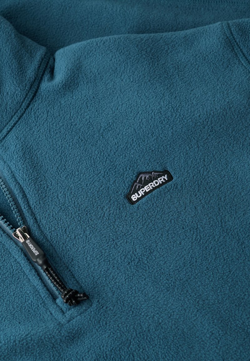 Teal fleece jacket met een halflange ritssluiting, een zachte textuur en een zwart geborduurd logo met een bergontwerp op de borst.