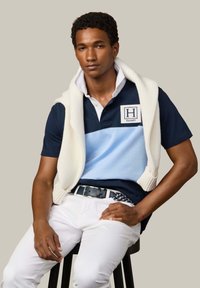 Jeune homme assis sur un tabouret portant un polo bleu marine et bleu clair avec un pull blanc enroulé autour des épaules, un pantalon blanc et une ceinture tressée.