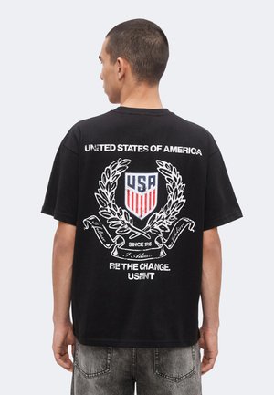 Homme portant un t-shirt noir avec un écusson en forme de bouclier des États-Unis, une couronne de laurier, et le texte "United States of America", "Be the Change" et "USMNT" au dos.