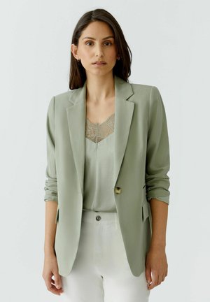 Frau mit langen dunklen Haaren, die einen salbeigrünen Blazer über einem spitzenbesetzten Top und weißen Hosen trägt, vor einem schlichten hellen Hintergrund stehend.