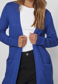 Cárdigan azul de punto con frente abierto, mangas largas, dos bolsillos delanteros y puños acanalados. Lleva puesto sobre una camiseta blanca y pantalones negros.