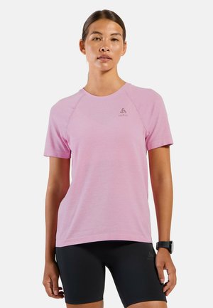 Jonge vrouw die een lichtroze sportshirt met korte mouwen en zwarte aansluitende shorts draagt, staand tegen een effen witte achtergrond.