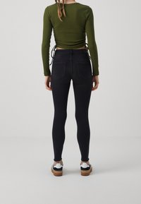 Pantalones negros de cintura alta con un ajuste elegante, hechos de denim elástico. Combinados con una blusa verde de manga larga, que presenta fruncidos laterales y lazos.