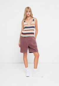 Gestreiftes gestricktes Top mit V-Ausschnitt in Pastellfarben; kombiniert mit lockeren, burgunderfarbenen Cordshorts; weiße Sneaker runden den Look ab.