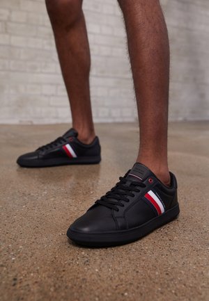 Personne portant des baskets noires avec des bandes rouges, blanches et bleues, debout sur un sol en béton poli contre un mur en briques clair.