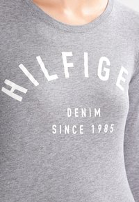 Grå bomullströja med fet vit text: "HILFIGER" bågformat ovanför "DENIM" och "SINCE 1985" i ett enkelt, modernt typsnitt.