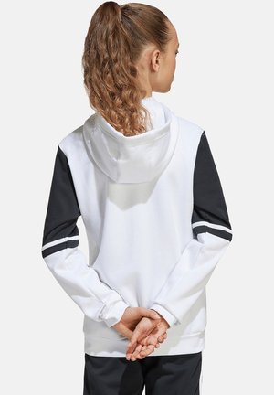 Junge Frau mit lockigem Pferdeschwanz, die einen weißen Hoodie mit schwarzen Ärmeln und Streifen trägt, die Hände hinter dem Rücken verschränkt, von hinten zu sehen.