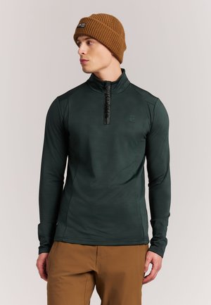 Jonge man die een donkergroen shirt met lange mouwen en rits draagt, bruine broek en een bruine gebreide muts, kijkt naar links.