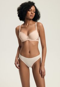 Kobieta z kręconymi włosami, ubrana w nagą bieliznę koronkową i kremowe majtki, stojąca na tle w jednolitym, neutralnym kolorze.