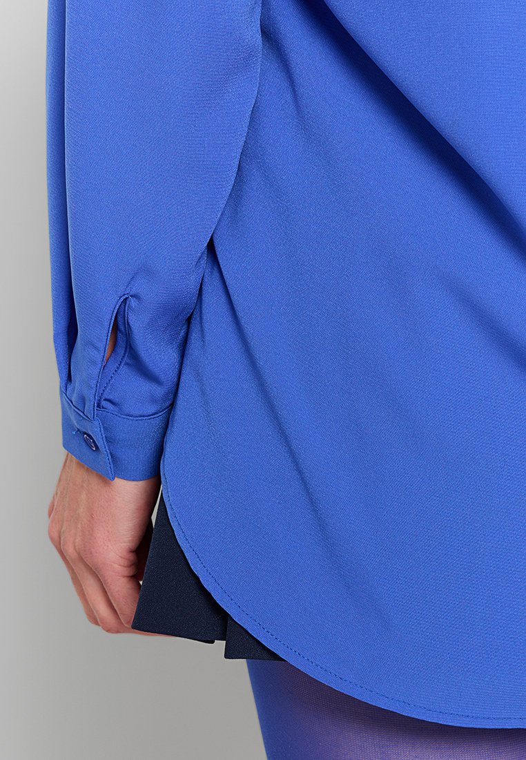 Blaues Langarmhemd mit glatter Textur, Knopfmanschetten und Seitenschlitzen. Der Saum ist leicht gebogen und hat ein geschichtetes schwarzes Detail darunter.