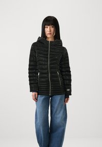 Svart isolert jakke med hette, med glidelåslommer, kviltet design og en slank finish. Sammen med løse blå jeans.