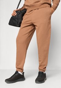 Brown jogger pants in soft material with an elastic waistband and cuffs. Paired with black mesh sneakers and a small black bag.
