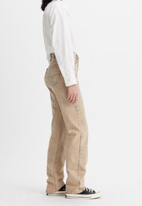 Jean en denim beige à coupe droite, dotée d'une taille haute, de coutures classiques et d'une texture subtile. Portée avec une chemise blanche à boutons et des baskets noires.