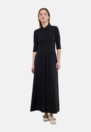 Femme portant une robe midi noire à col boutonné avec des manches trois quarts et des chaussures Mary Jane beiges, debout devant un fond blanc.