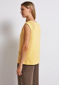 Top senza maniche giallo in tessuto testurizzato, con dettaglio in pizzo sulla schiena. Abbinato a pantaloni larghi in lino marrone.