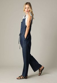 Denim overall met een fitted ontwerp, donkerblauwe kleur, wijde pijpen, een ceintuur en een voorzak. Gecombineerd met een witte top met kant en sandalen.