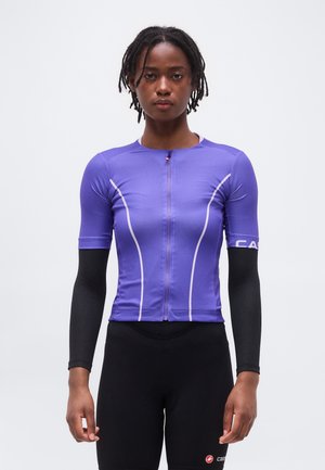 Jong persoon met korte dreadlocks die een nauwsluitend paars wielershirt draagt met zwarte armverwarmers en zwarte wielershorts.