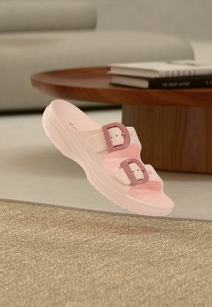 Sandalia slip-on rosa claro con dos hebillas ajustables de color rosa más oscuro que flota sobre una alfombra beige tejida cerca de una mesa de café de madera.