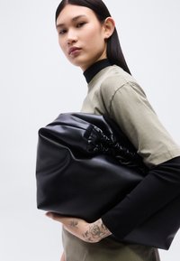 Νεαρό άτομο με μακριά σκούρα μαλλιά κρατά μια μεγάλη μαύρη τσάντα από μαλακό δέρμα, φορώντας ελιάς πράσινο πουκάμισο πάνω από μαύρα μακριά μανίκια, δείχνοντας το τατουάζ στο χέρι.