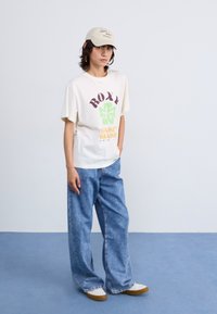 Cream t-shirt with Roxy logo and colorful graphics, paired with loose blue jeans and beige sneakers, set against a light background.