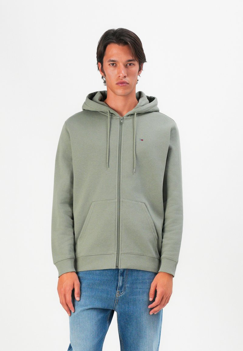 Hombre vestido con una sudadera verde con cremallera y un pequeño logo en el pecho, con jeans azules, de pie contra un fondo blanco plano.