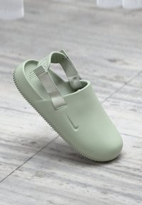 Ανοιχτό πράσινο σανδάλι χωρίς κορδόνια με ρυθμιζόμενους υφασμάτινους ιμάντες και υφή στη σόλα, τοποθετημένο σε δάπεδο με ανοιχτό γκρι πλακάκια.