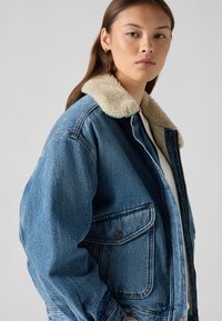 Veste en denim avec col en fausse fourrure, couleur bleue, coupe ample, poche avant et détails de couture visibles. Le tissu texturé offre une apparence chaleureuse.