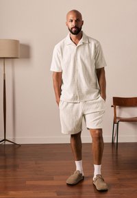 Camicia a maniche corte a righe chiare e pantaloncini abbinati in colori pallidi, realizzati in tessuto strutturato. Abbinati a scarpe slip-on beige e calzini bianchi.