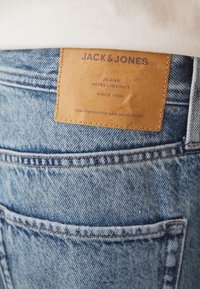 Jean en denim bleu avec une étiquette en cuir marron sur la poche arrière, présentant un texte en relief : "JACK & JONES, JEANS INTELLIGENCE, SINCE 1990."