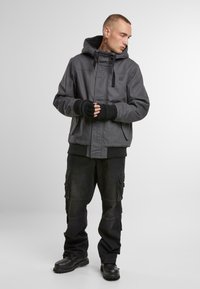 Pilkas gobtuvas striukė su priekinėmis kišenėmis, juodi raibalių rankogaliai ir juodos krovininės kelnės. Avalynė - juodi batai su tekstūruotu padu.