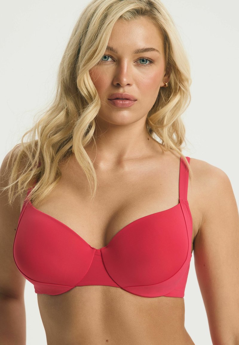 Blondynka o falowanych włosach nosi czerwony biustonosz z fiszbinami na jasnym, jednolitym tle, zwrócona przodem z neutralnym wyrazem twarzy.