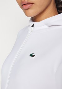 Hvit zip-up hettegenser laget av glatt stoff, med en liten grønn krokodille-logo med rød aksent på venstre brystområde.